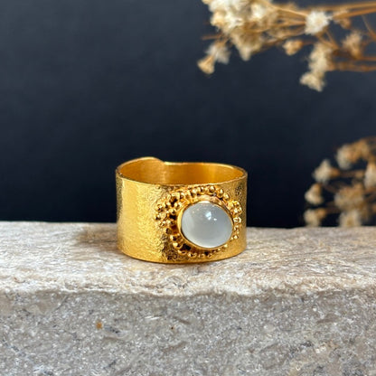 A wide, textured gold ring with a central oval white stone, displayed on a light gray stone surface against a dark background with dried flowers.