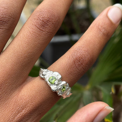 A close-up of a hand wearing a textured silver ring with two small green gemstones, set against a blurred background of green foliage.
