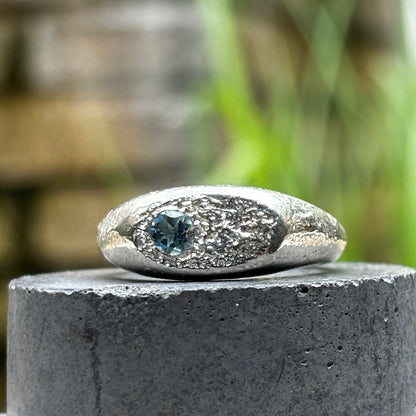 A silver ring with a textured, organic-shaped setting and a central round blue gemstone, displayed on a dark stone pedestal with a blurred outdoor background.