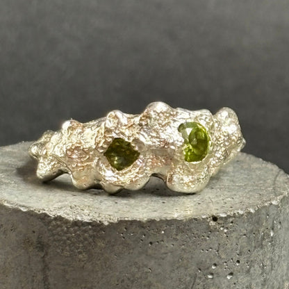 A textured sterling silver ring with two oval green gemstones, displayed on a cylindrical gray concrete pedestal against a dark gray background.