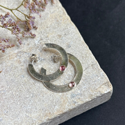 Two silver crescent-shaped hoop earrings with small pink gemstone accents, displayed on a textured light gray stone slab against a dark background.