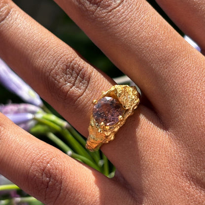 A close-up of a hand wearing a textured gold ring with a round purple gemstone, set against a blurred background of green leaves and purple flowers.