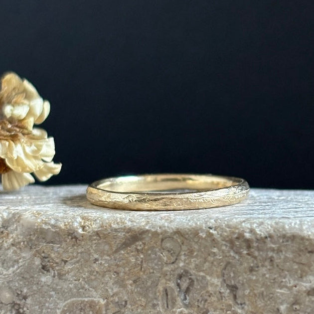 A textured gold band ring resting on a rough stone surface next to dried cream-colored flowers against a dark background.