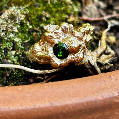 A close-up of a gold-toned ring with a textured, organic shape and a central dark green faceted gemstone, resting on moss and soil next to a terracotta pot.