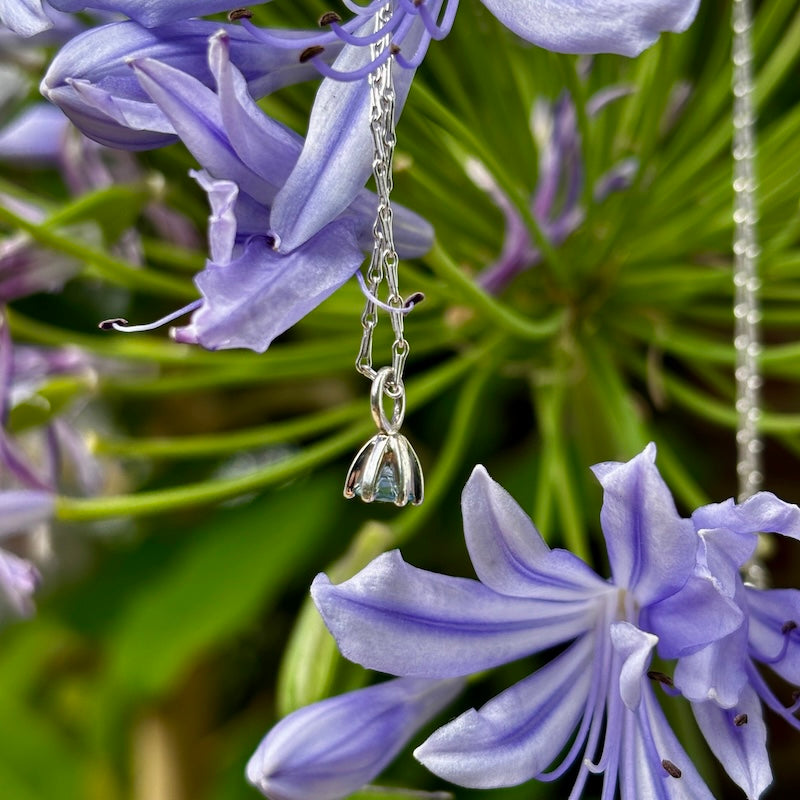 A silver-toned pendant necklace with a small hanging flower charm is photographed outdoors among purple flowers and green foliage.