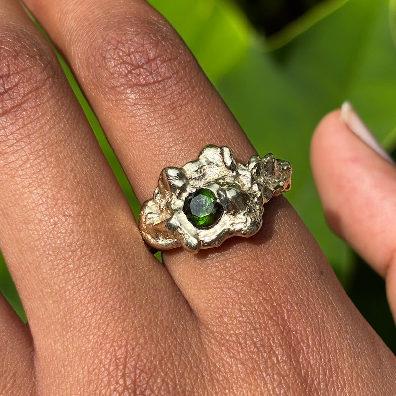A close-up of a textured silver ring with a central green gemstone, worn on a person's finger against a blurred green background.
