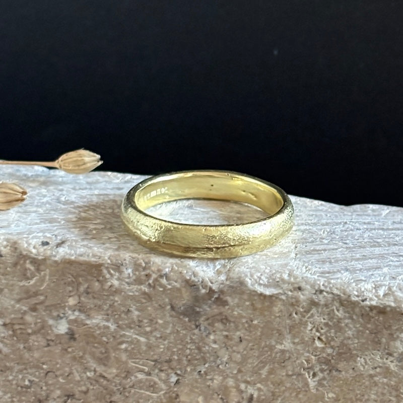 A textured yellow gold band ring resting on a light-colored stone slab against a dark black background.