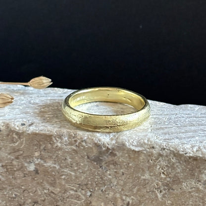 A textured yellow gold band ring resting on a light-colored stone slab against a dark black background.
