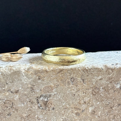 A textured yellow gold band ring resting on a rough stone surface against a solid black background, with small dried flower buds as a decorative prop on the left.