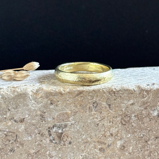 A textured yellow gold band ring resting on a rough stone surface against a solid black background, with small dried flower buds as a decorative prop on the left.