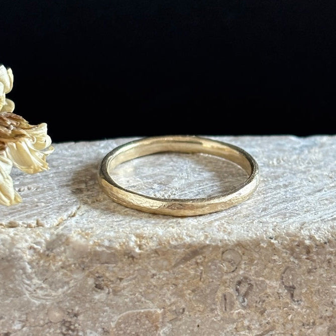 A textured yellow gold band ring resting on a light beige stone surface next to dried flowers, with a solid black background behind.