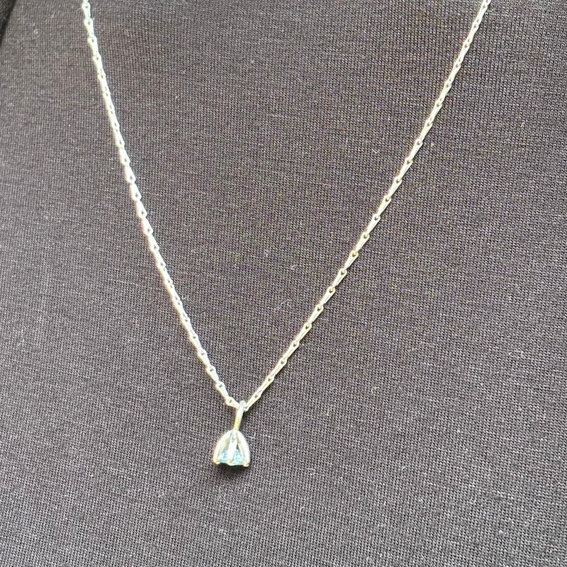 A delicate silver chain necklace with a small teardrop-shaped pendant featuring a light blue iridescent stone, displayed on a textured dark gray fabric background.