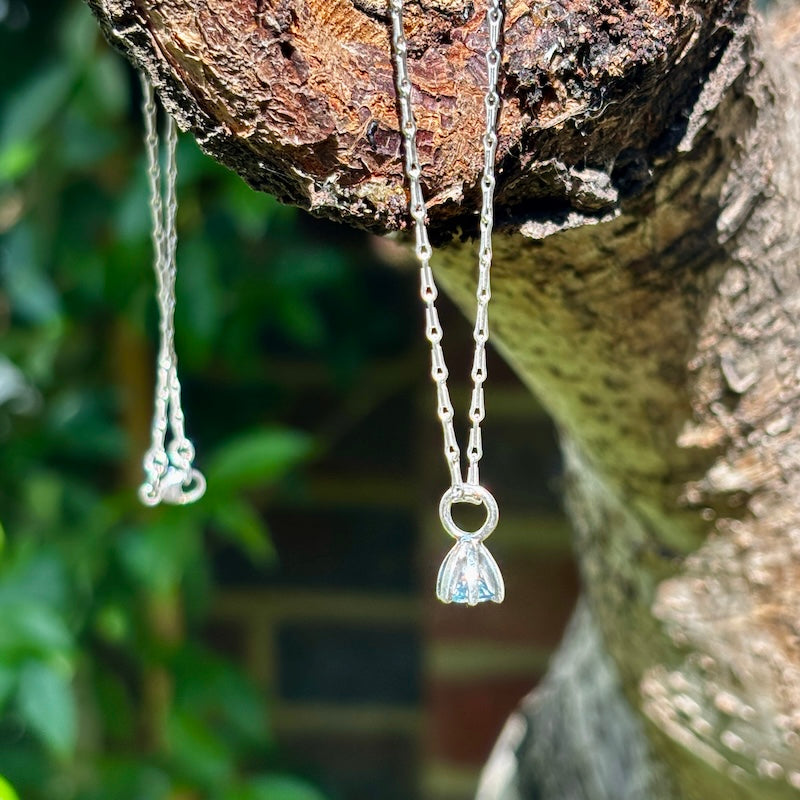 A silver-toned necklace with a small floral pendant hangs from a tree branch outdoors, with a blurred green garden background.