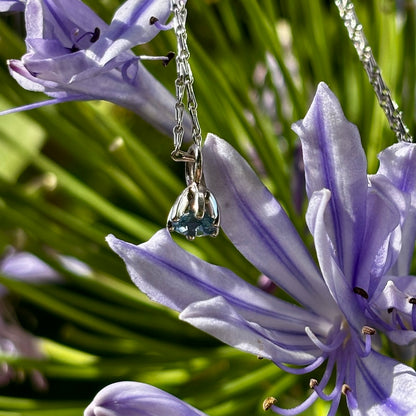 A silver chain necklace with a small clear teardrop-shaped crystal pendant, photographed close up against purple flowers and green foliage.