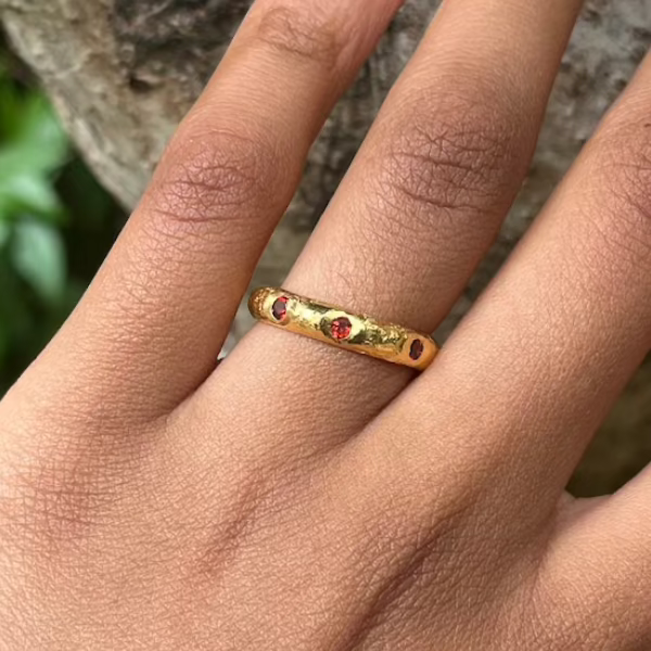 A gold band ring with small red gemstone accents, shown worn on a person's finger against a blurred outdoor background.