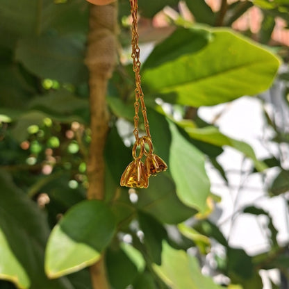 A gold-toned metal chain with three small bell-shaped charms, photographed hanging against a background of green leaves and a tree trunk.