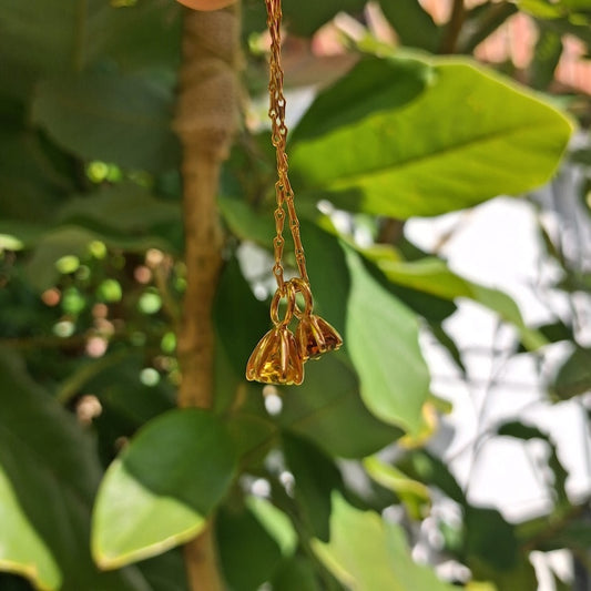 A gold-toned metal chain with three small bell-shaped charms, photographed hanging against a background of green leaves and a tree trunk.