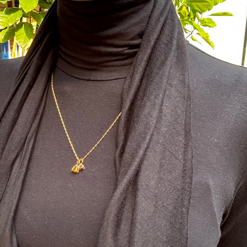 A gold chain necklace with a small butterfly pendant is worn by a person wearing a black turtleneck and a black scarf, with green leaves visible in the background.