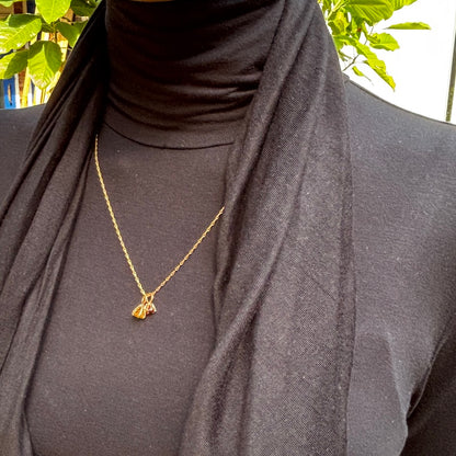 A gold chain necklace with a small butterfly pendant is worn by a person wearing a black turtleneck and a black scarf, with green leaves visible in the background.