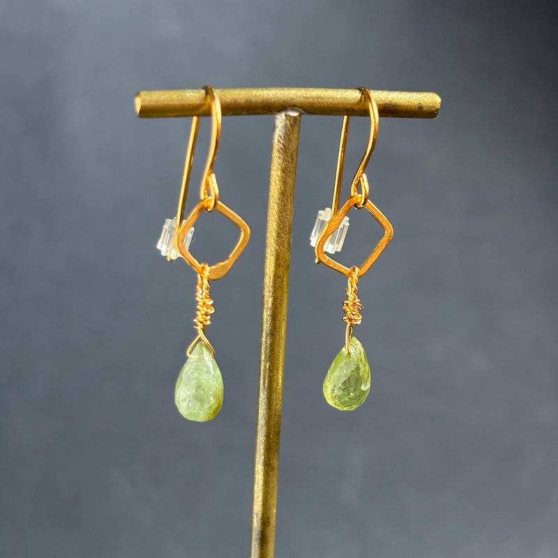A pair of gold drop earrings with geometric wireframe loops and faceted light green teardrop gemstones, displayed on a gold stand against a dark gray background.