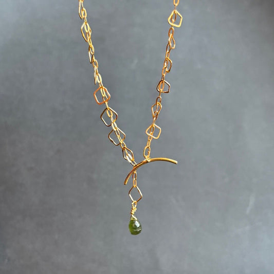 A delicate gold chain necklace with geometric links and a small green teardrop gemstone pendant, shown against a dark gray textured background.