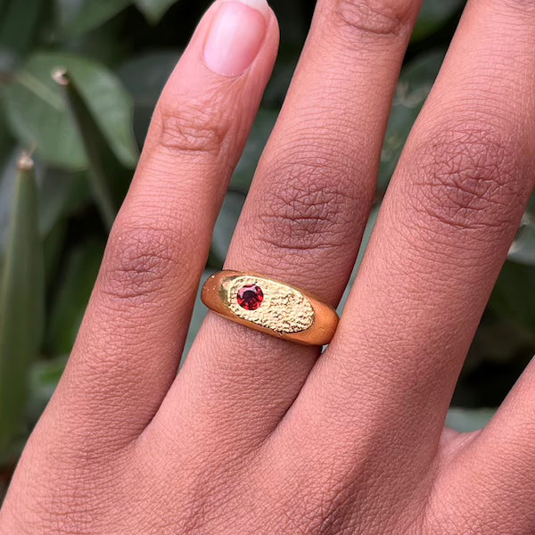 A gold ring with a textured oval band and a small red gemstone in the center, worn on a person's finger with a blurred green background.
