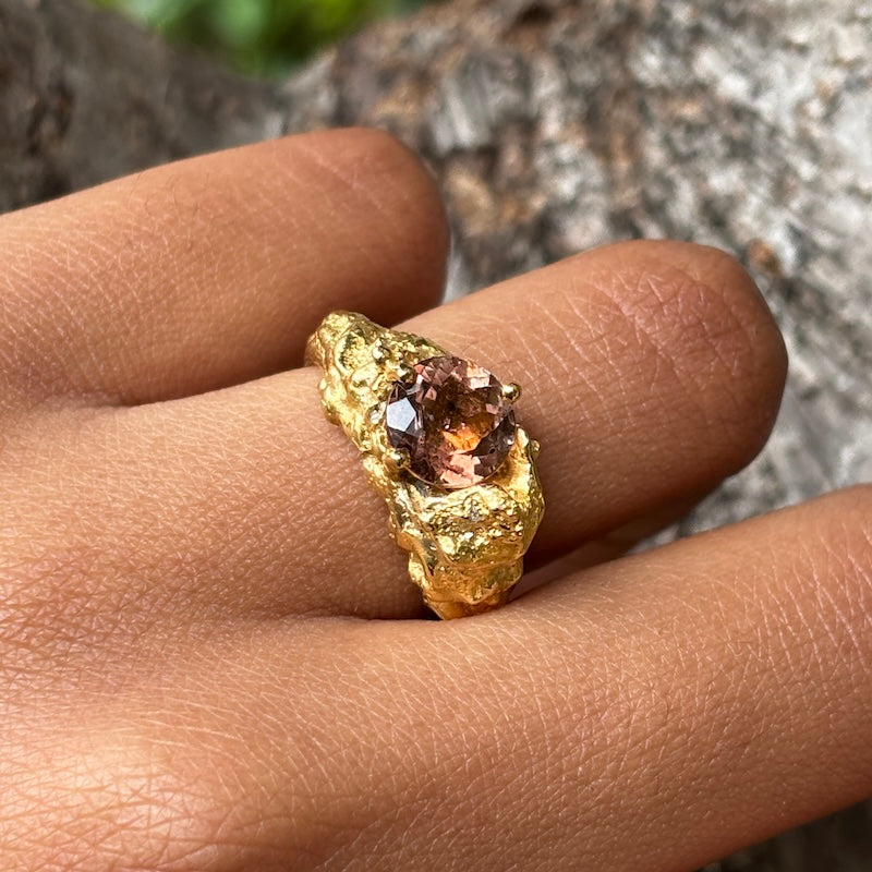 A close-up of a hand wearing a textured gold ring with a faceted amber-brown gemstone, set against a blurred natural background.