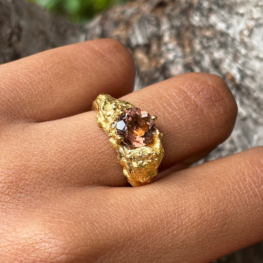 A close-up of a hand wearing a textured gold ring with a faceted amber-brown gemstone, set against a blurred natural background.