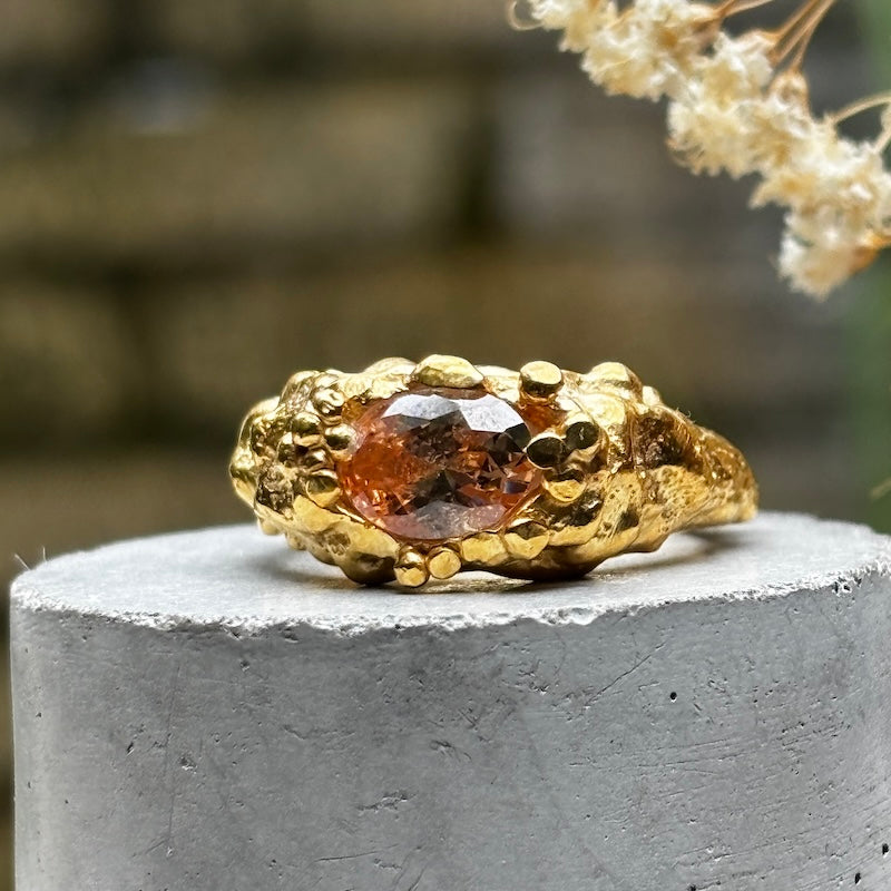 A textured gold ring with a central faceted amber-brown gemstone, displayed on a round gray concrete pedestal with a blurred natural background.