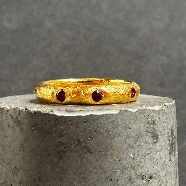 A textured gold ring with three small round red gemstones, displayed on a gray cylindrical pedestal against a gray background.