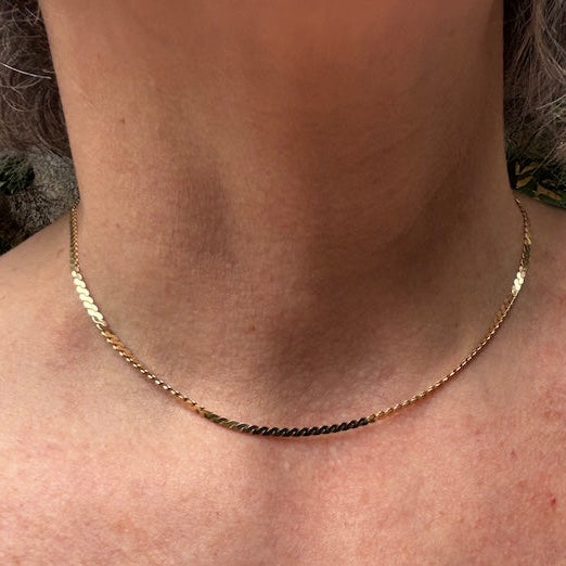 A close-up of a person's neck and upper chest wearing a delicate gold chain necklace. The necklace has a textured, diamond-like pattern along the chain.