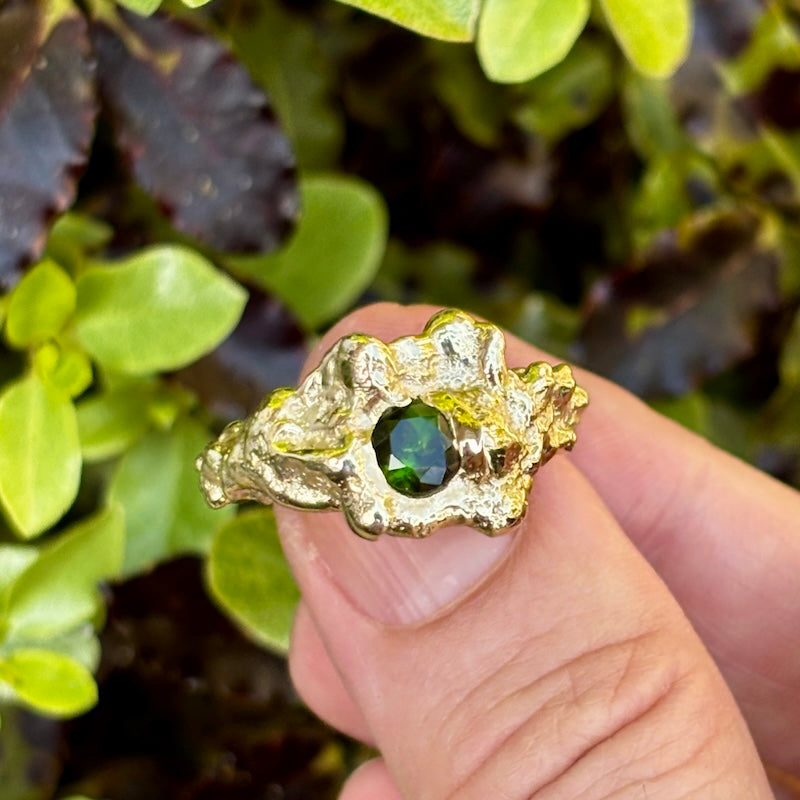 A hand holding a gold ring with a textured, organic shape and a central green faceted gemstone, set against a background of green foliage.