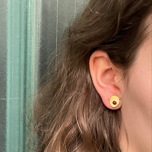 Close-up of an ear wearing a gold earring with a red gemstone against a blurred background